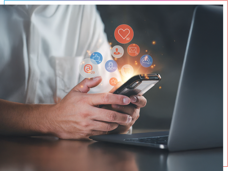 Person sitzt am Tisch vor dem Laptop und Smartphone. Icons schweben über dem Bildschirm.