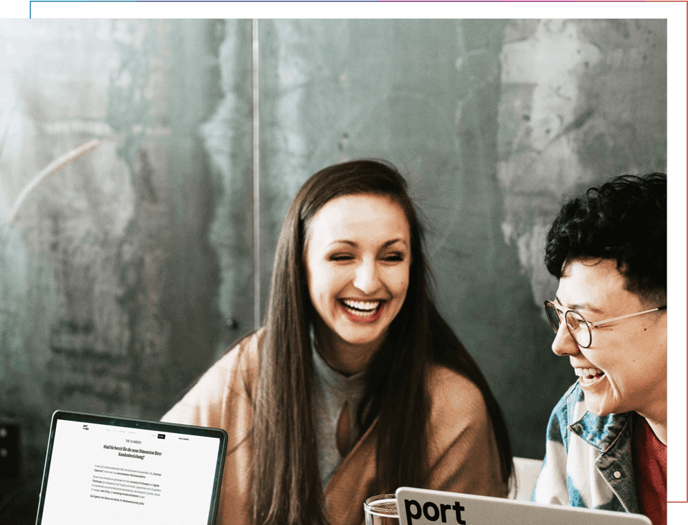 Zwei Kolleginnen lachen gemeinsam bei der Arbeit an ihren Laptops.