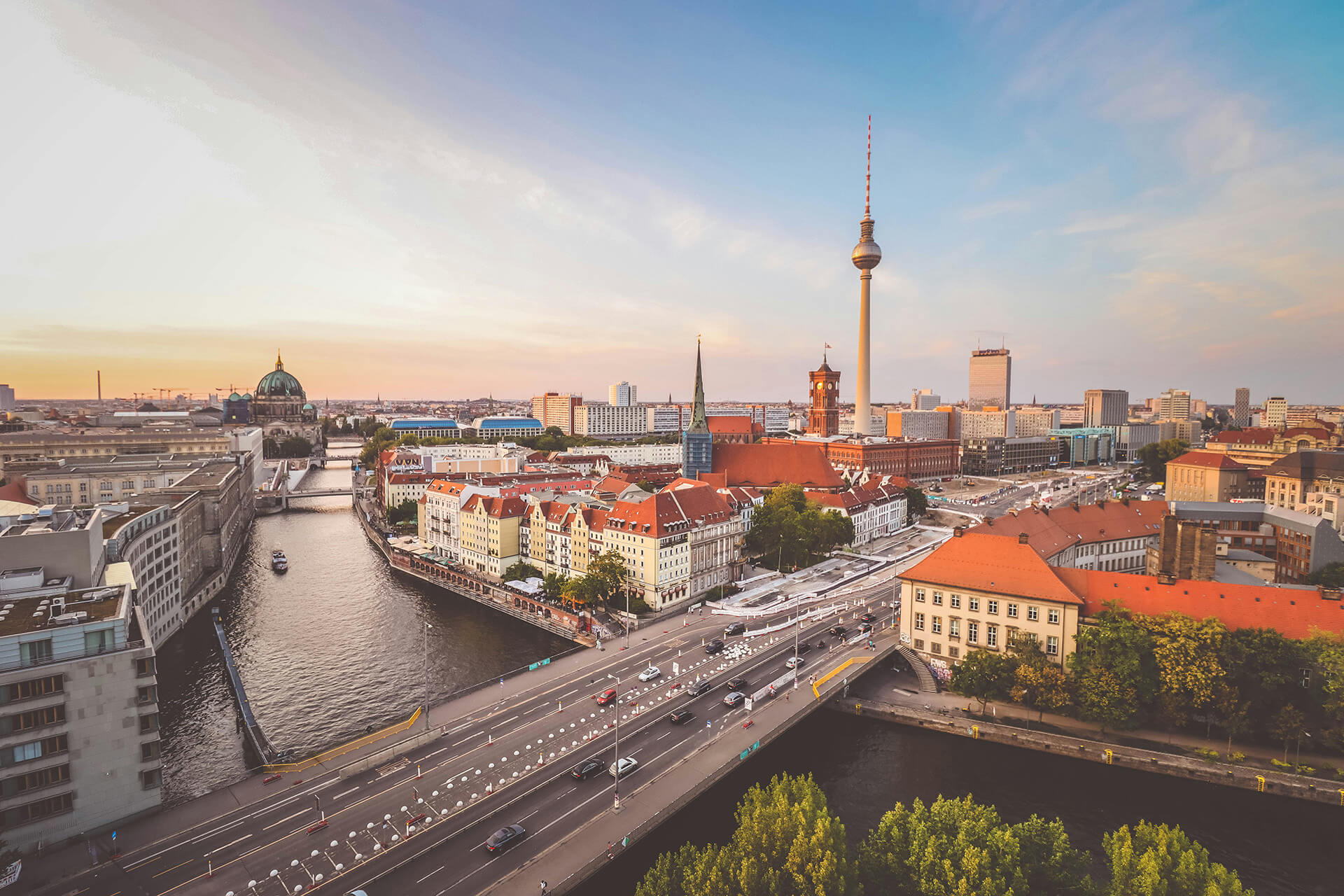 Panoramablick über Berlin bei Sonnenuntergang mit der Spree, historischen Gebäuden, roten Dächern und dem Fernsehturm im Hintergrund.