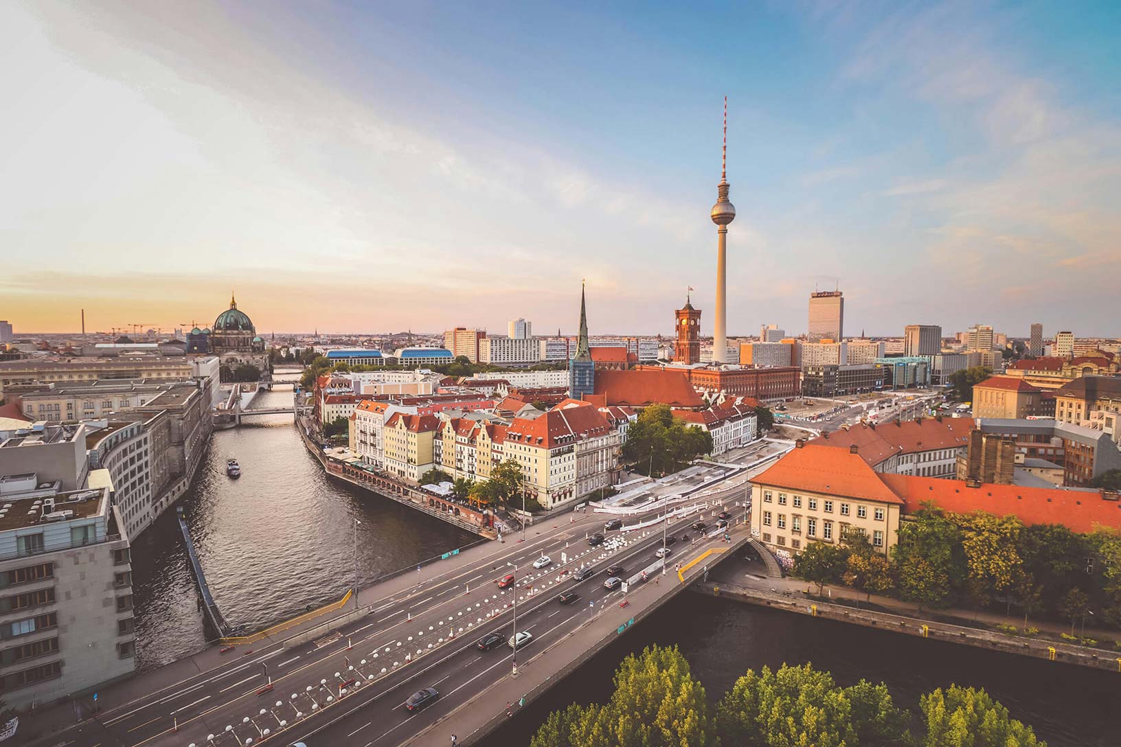 Panoramablick über Berlin bei Sonnenuntergang mit der Spree, historischen Gebäuden, roten Dächern und dem Fernsehturm im Hintergrund.