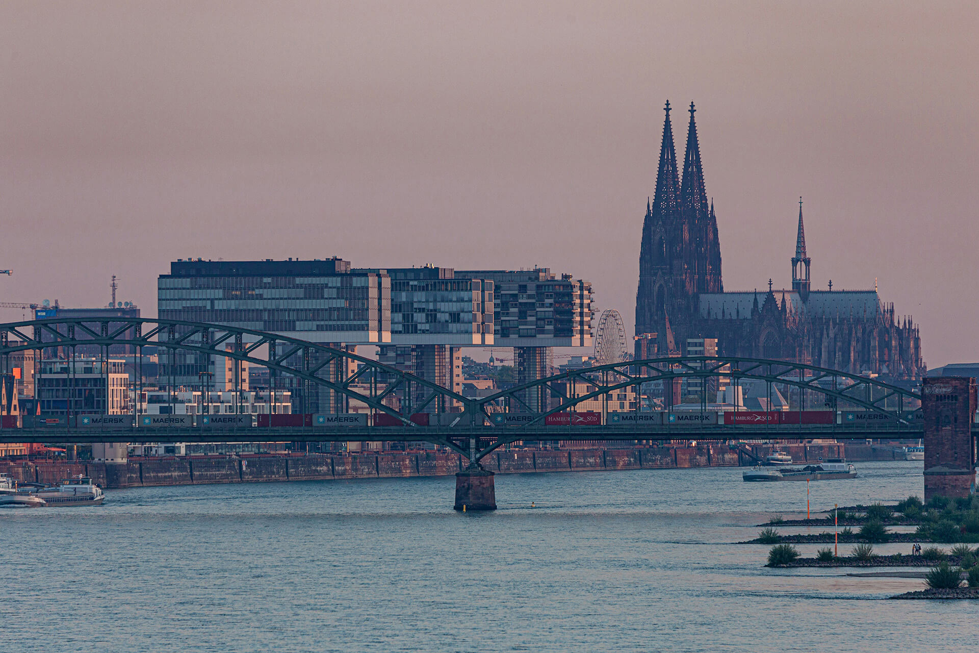 Blick über den Rhein auf die Hohenzollernbrücke und den Kölner Dom in der Abenddämmerung.