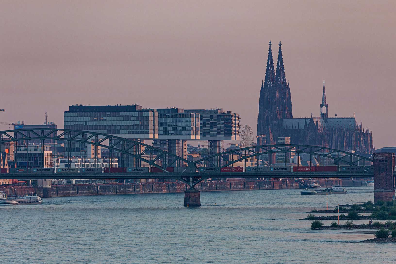 Blick über den Rhein auf die Hohenzollernbrücke und den Kölner Dom in der Abenddämmerung.