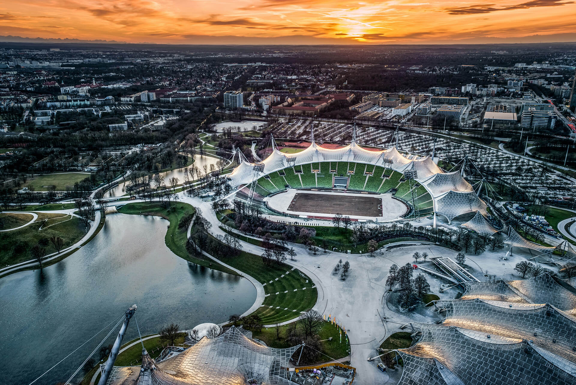 Luftaufnahme des Olympiastadions in München bei Sonnenuntergang mit Olympiapark, See und Zeltdach-Konstruktion.