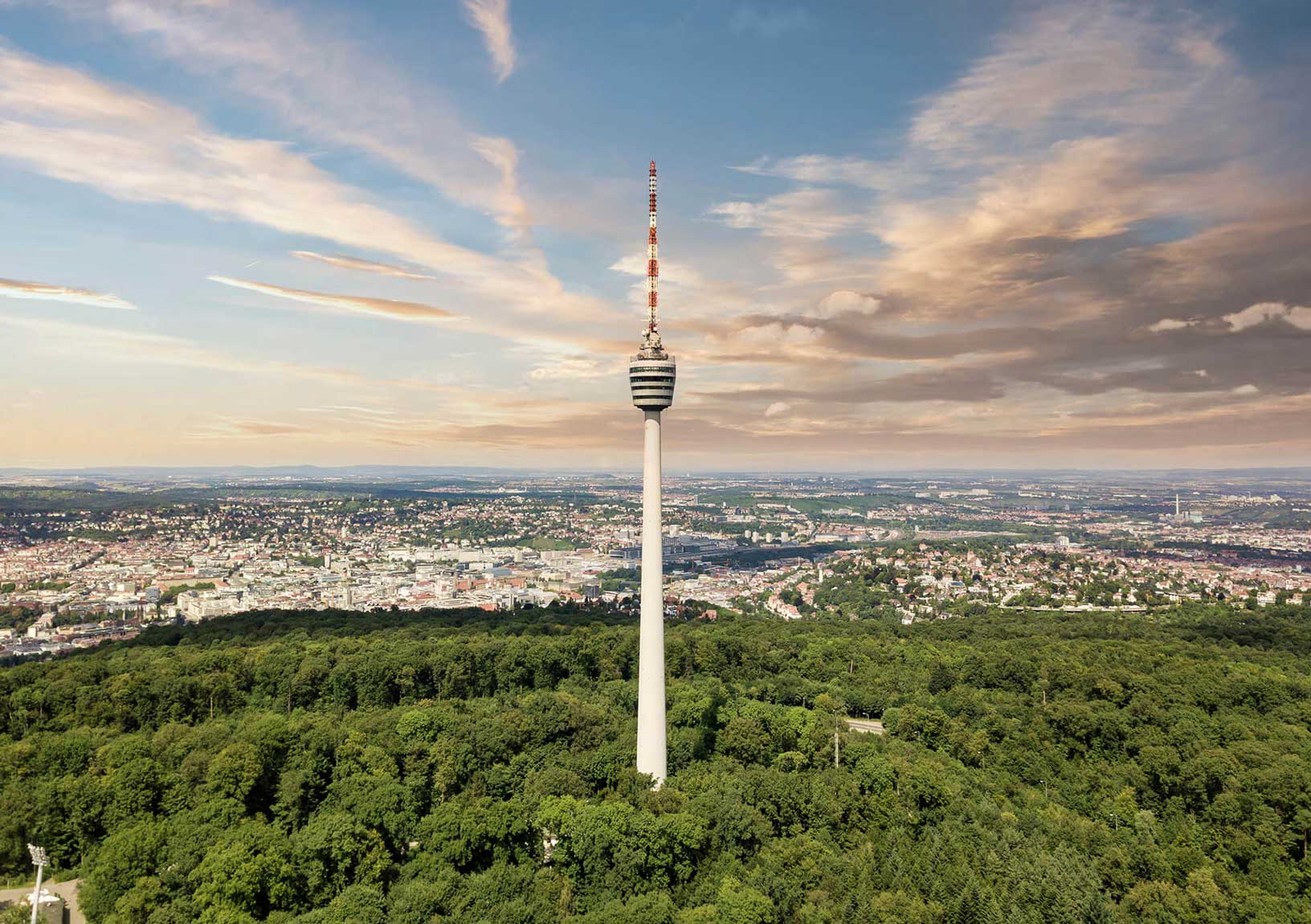 Der Stuttgarter Fernsehturm erhebt sich über einem dichten Wald, im Hintergrund ist die Stadt Stuttgart zu sehen.